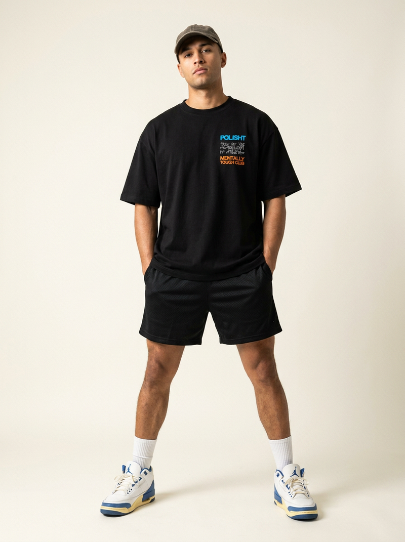 Man wearing a black t-shirt with text and graphics, black shorts, and a cap on a plain background