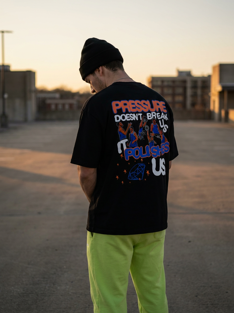 Person wearing a black t-shirt with colorful text and graphics, standing in an open area with buildings in the background.