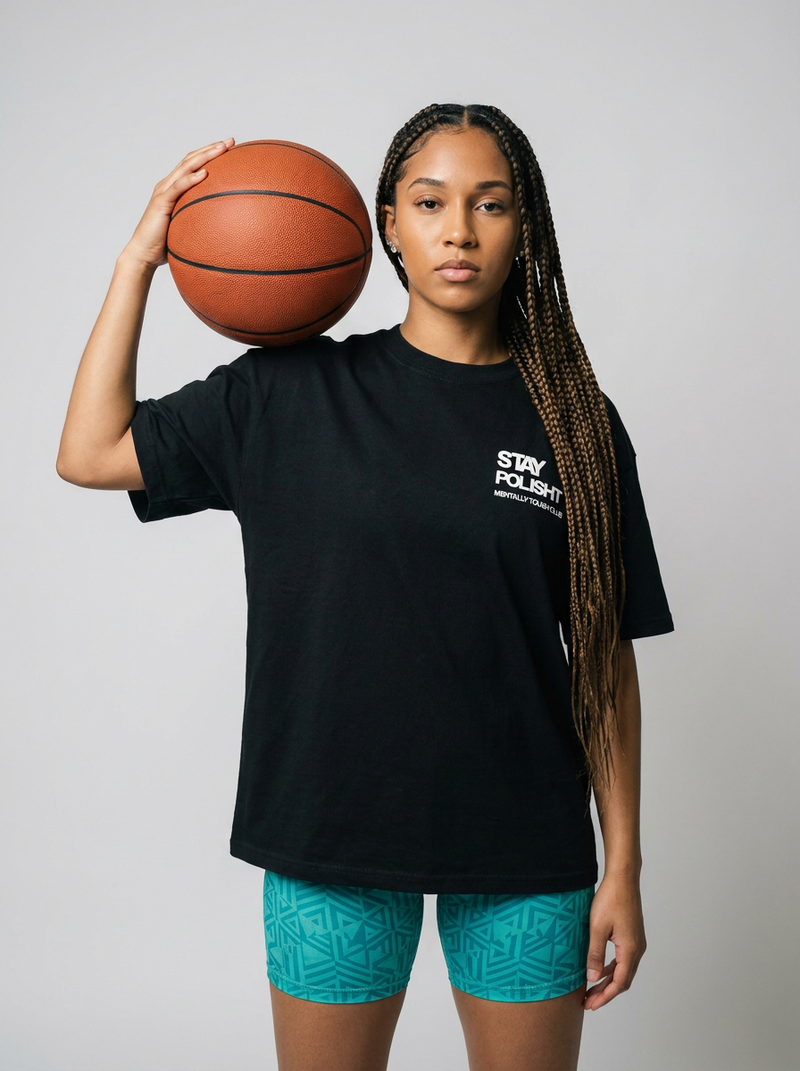 Feemale holding a basketball wearing a black t-shirt with text and teal shorts on a plain background